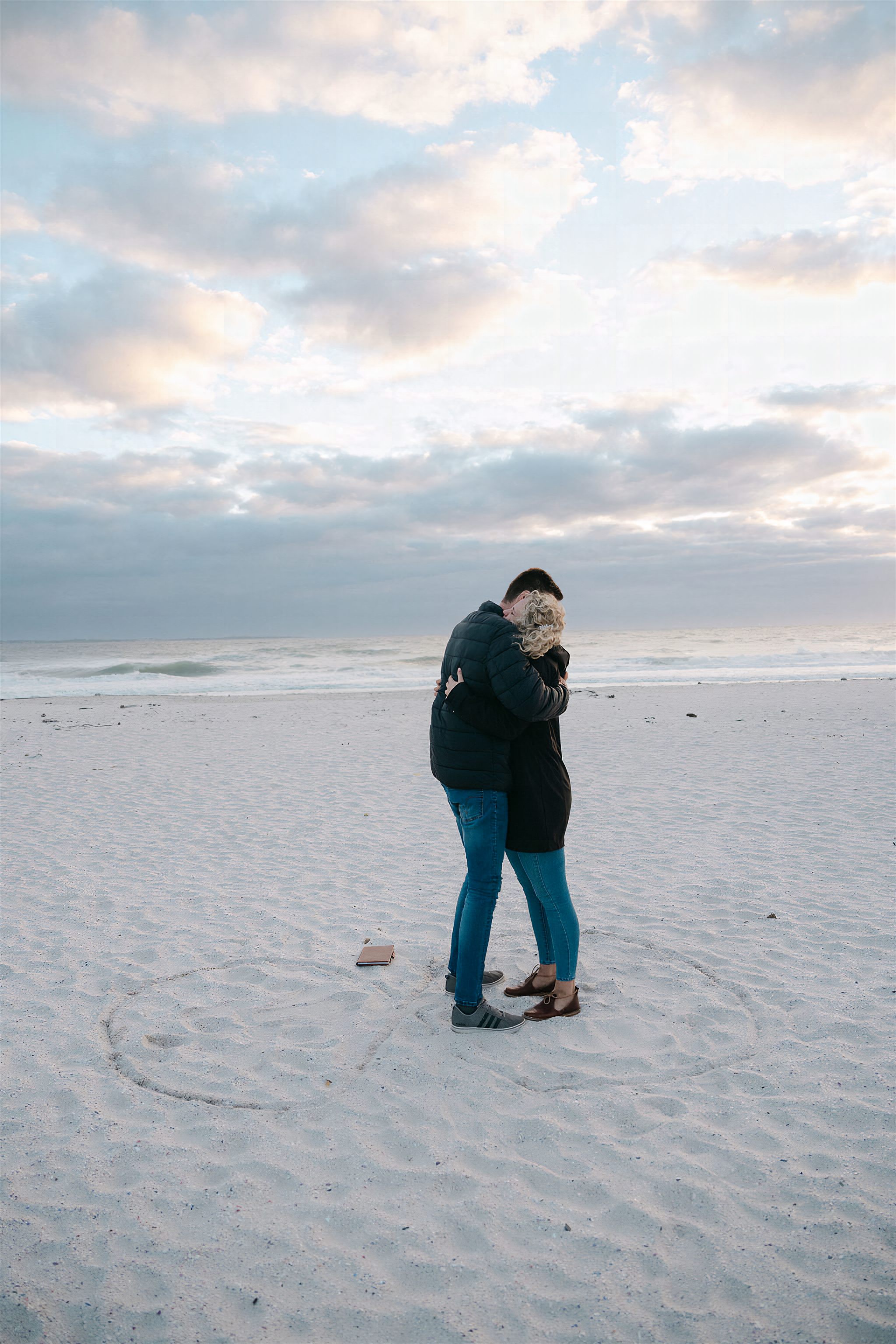 Kobus en Elrine omhels op die strand, 'n hart in die sand om hulle getrek